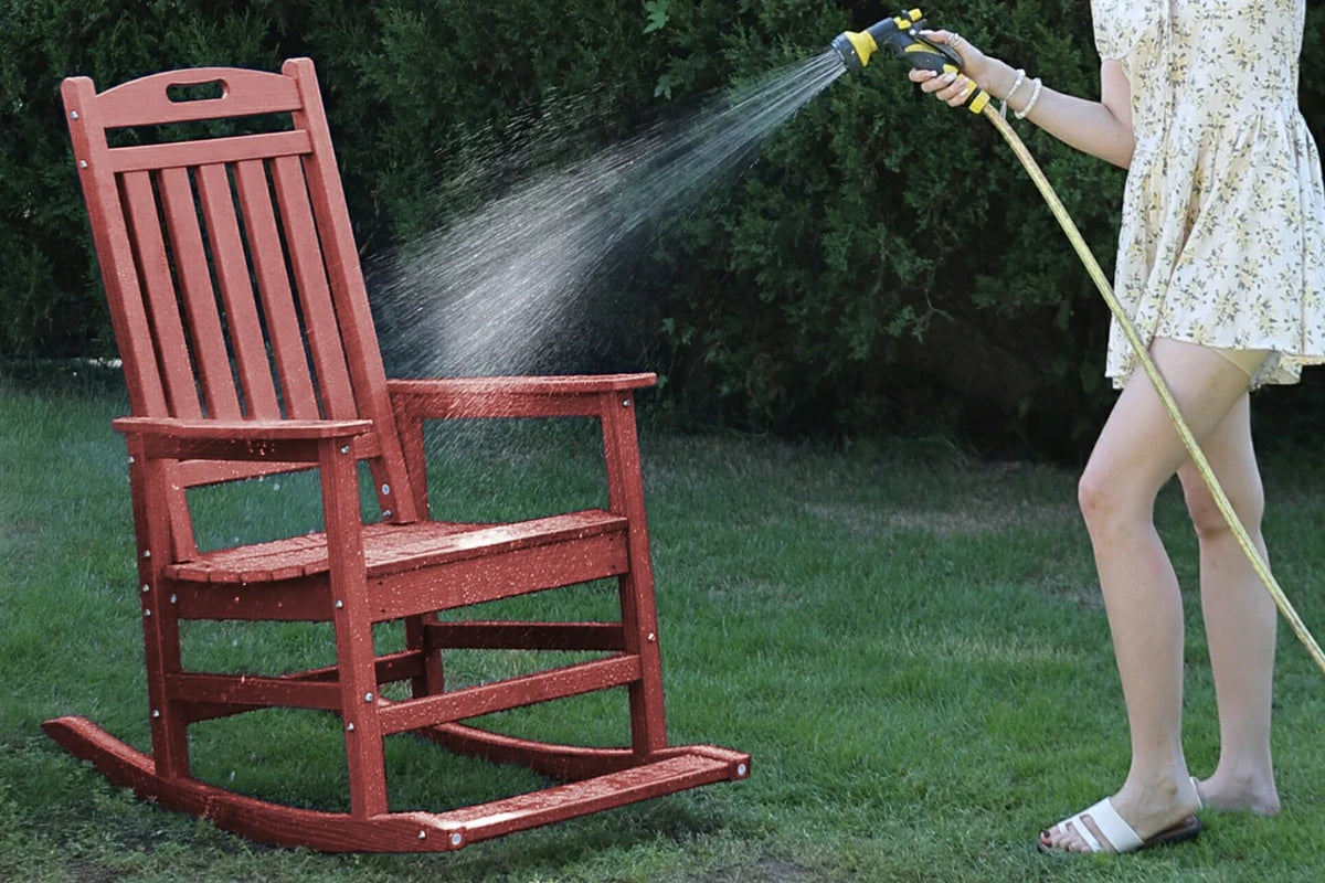 Red rocking chair being rinsed with a hose by a person on a grassy area.