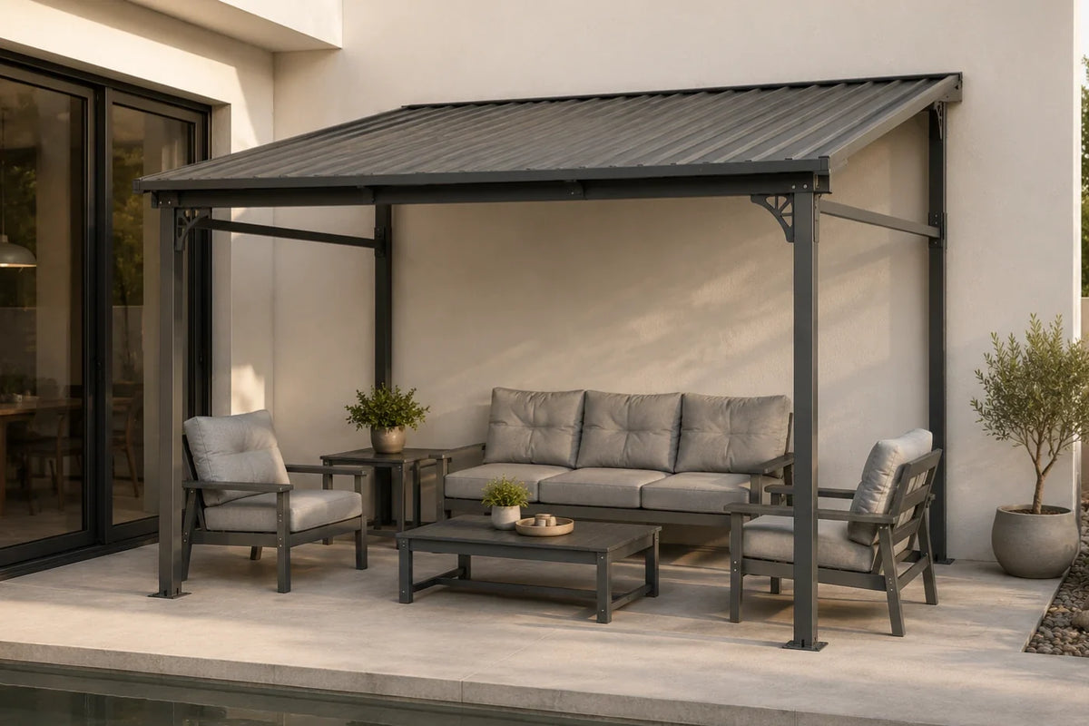 A modern outdoor patio setup featuring a dark gray wall-mounted hardtop gazebo, sheltering a gray cushioned three-seat sofa, two matching armchairs, and a low rectangular coffee table. The patio is set against a clean beige stucco wall next to a glass sliding door, with potted plants on either side.