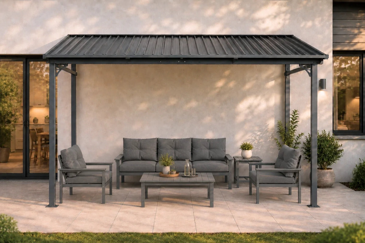 A dark gray wall-mounted hardtop gazebo, sheltering a four-piece outdoor conversation set on a light stone patio in front of a beige stucco house.