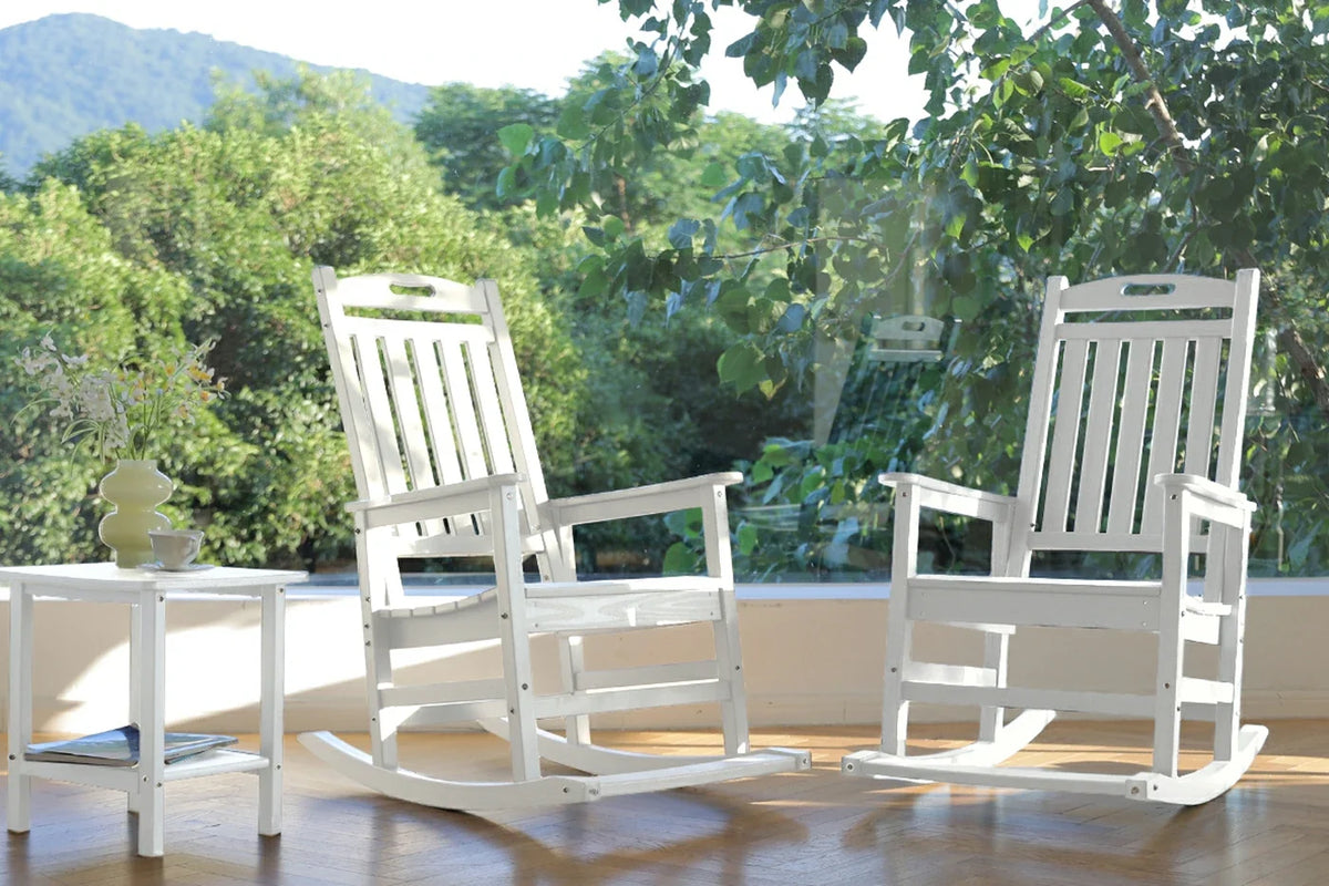 Two white wooden rocking chairs on a wooden floor with a green outdoor background.