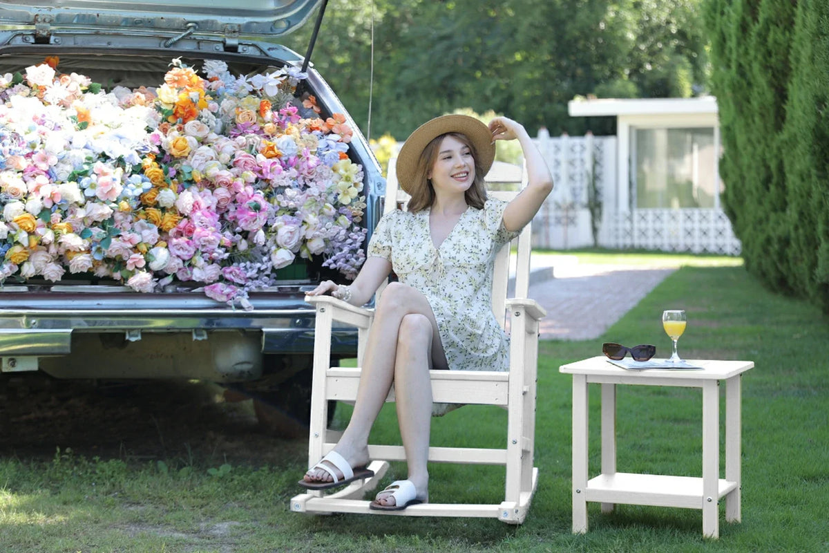 Woman sitting on a white chair next to a car filled with flowers, with a small table and sunglasses in the background.