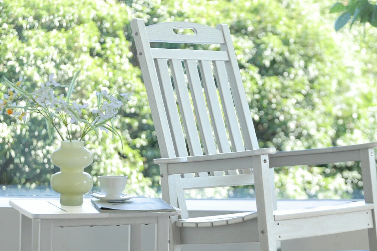 White rocking chair with a small table and decorative items on a blurred green outdoor background
