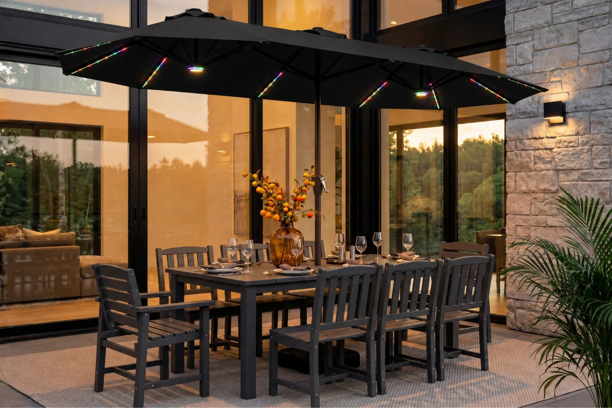 A large black patio umbrella with integrated warm white string lights is shown over a dark dining table and chairs on a patio. The setting is a modern home exterior at dusk with stone walls, glass windows, and a potted plant in the foreground.