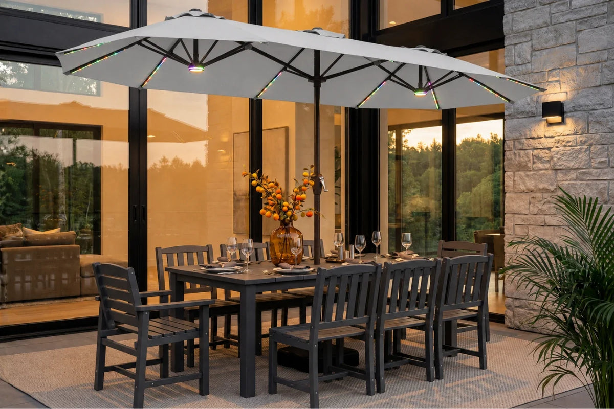 An outdoor patio dining set with a rectangular dark gray table and 8 matching slatted chairs, under a large gray patio umbrella with integrated lighting, set in front of a modern glass house.
