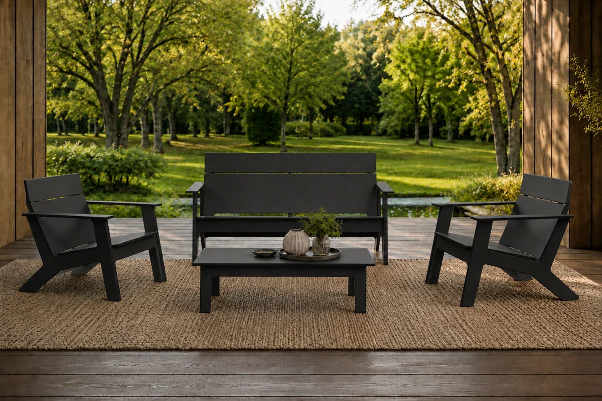 A 4-piece outdoor patio furniture set including a loveseat, two armchairs, and a coffee table, all in a black finish, arranged on a woven jute rug on a wooden porch with a lush green garden background.