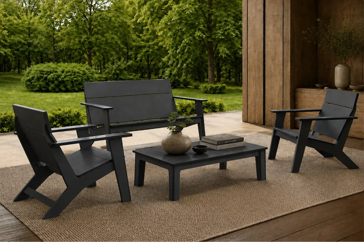 A 4-piece outdoor patio furniture set in black, including a loveseat, two armchairs, and a rectangular coffee table, arranged on a jute rug in a patio setting with a green garden background.