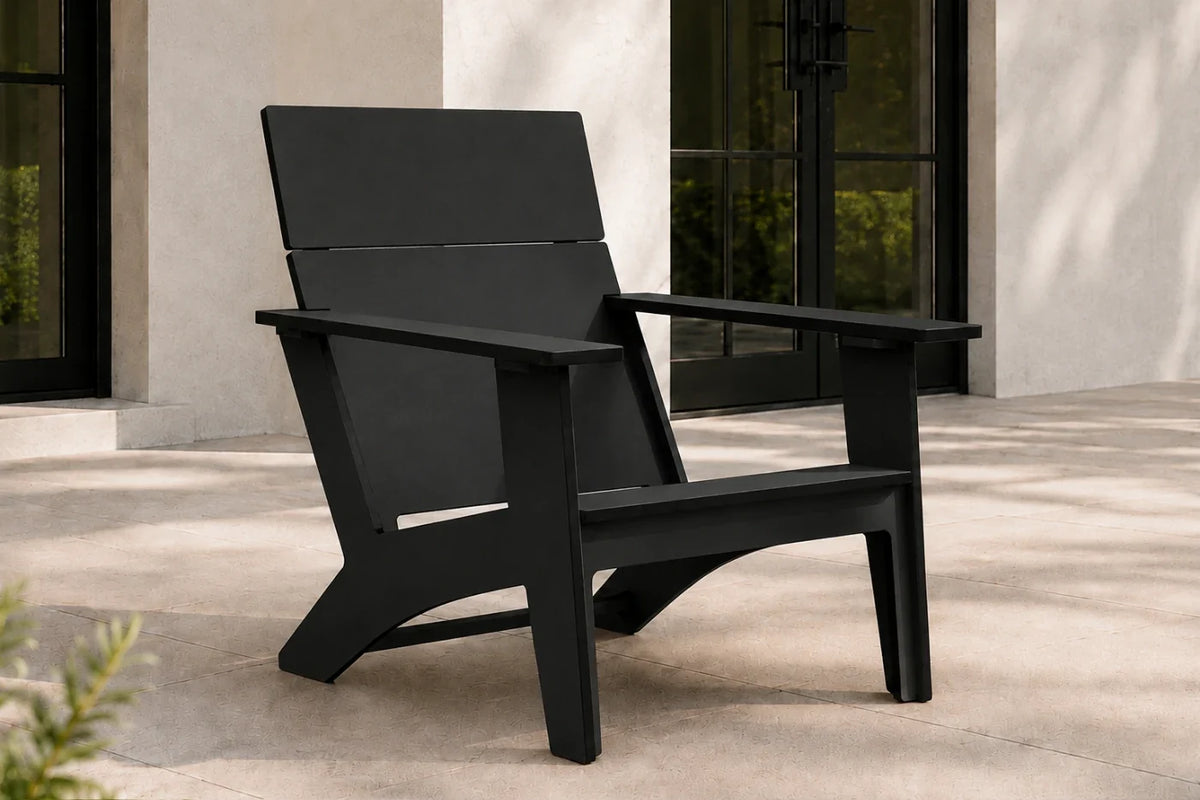 A modern black outdoor Adirondack chair with a wide back and armrests, positioned on a light stone patio in front of a building with dark-framed glass doors.