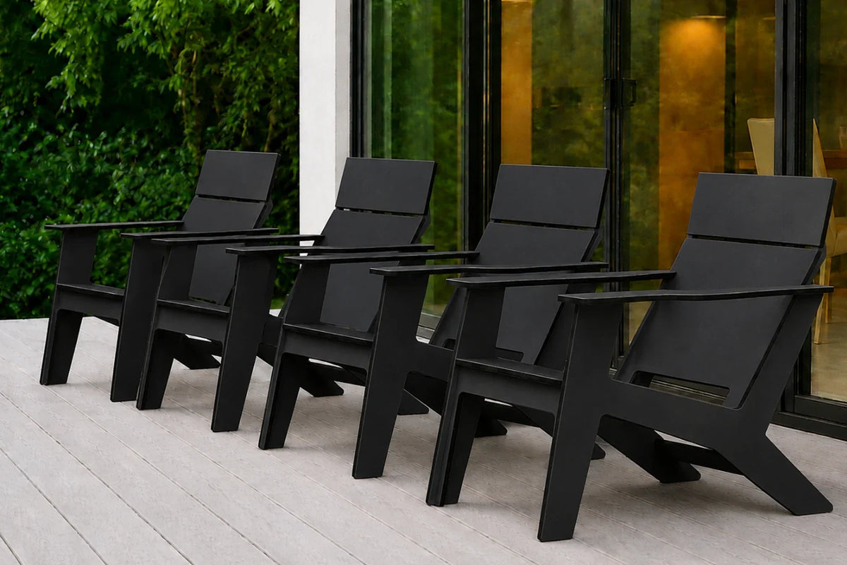 Four modern black outdoor Adirondack chairs arranged in a row on a light gray deck in front of a glass-walled building with greenery visible on the left.