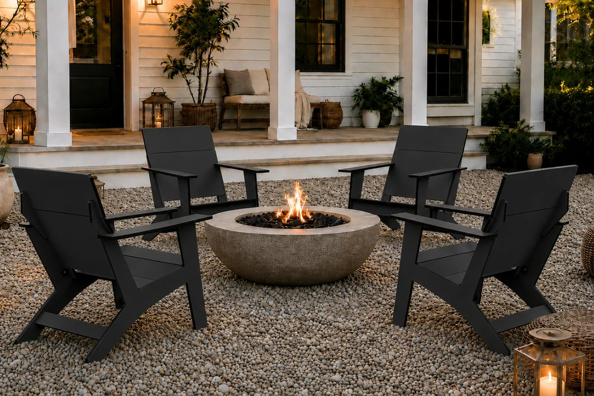 Four black outdoor Adirondack chairs arranged around a round concrete fire pit on a gravel patio in front of a white house porch at dusk.