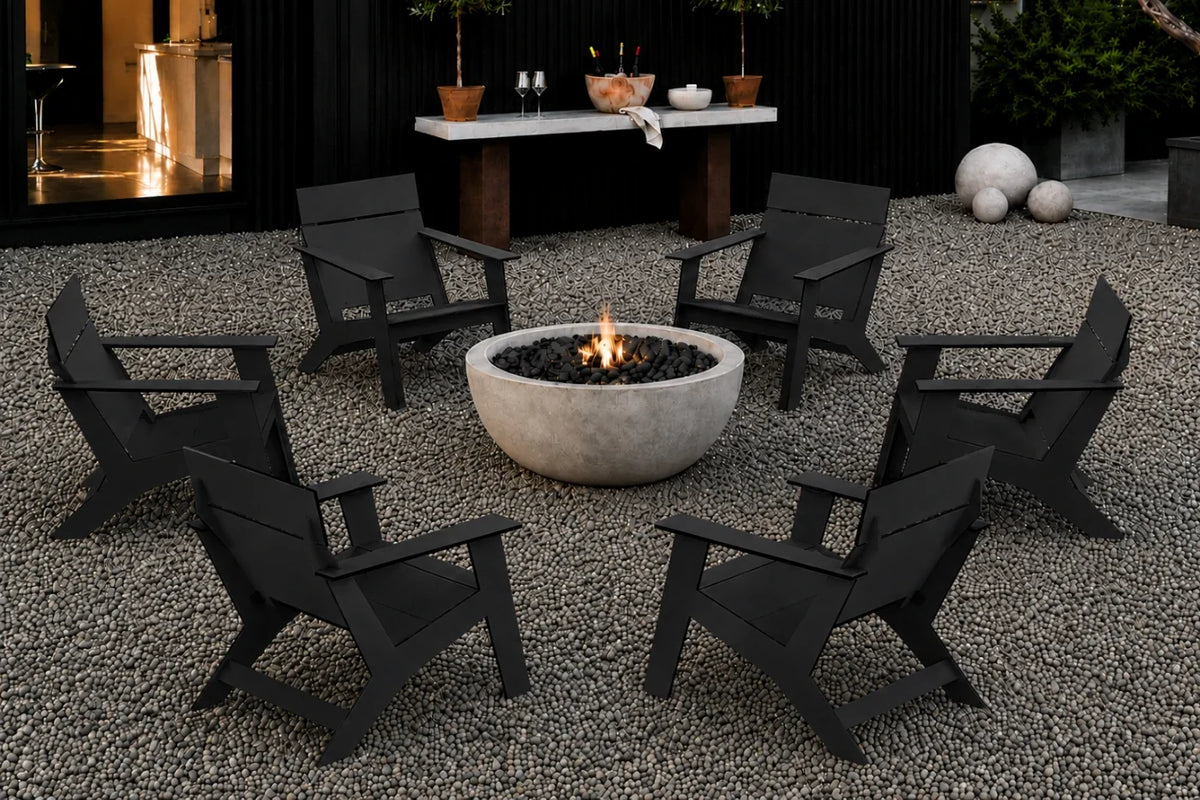 Six black outdoor Adirondack-style chairs arranged around a round concrete fire pit on a gravel patio, with a modern black wall and small table in the background.
