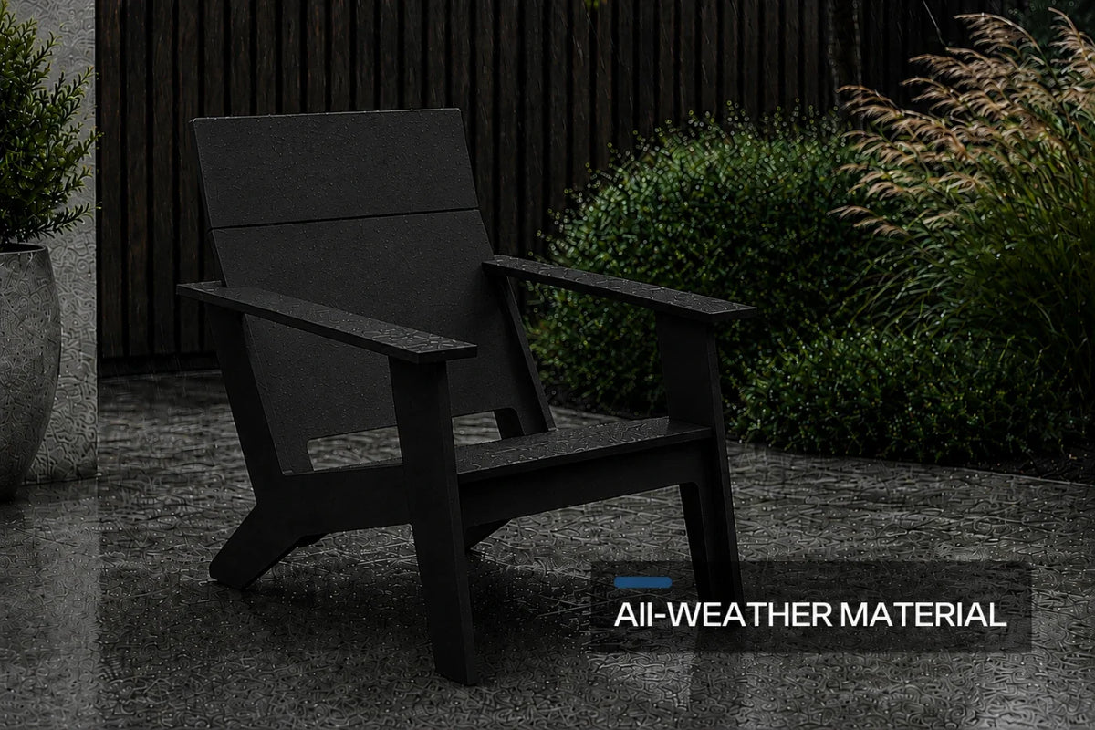 A black modern poly adirondack chair with a wide armrest and angled backrest, shown on a stone patio with shrubs and a dark fence in the background. A text overlay in the bottom right reads 'All-WEATHER MATERIAL'.