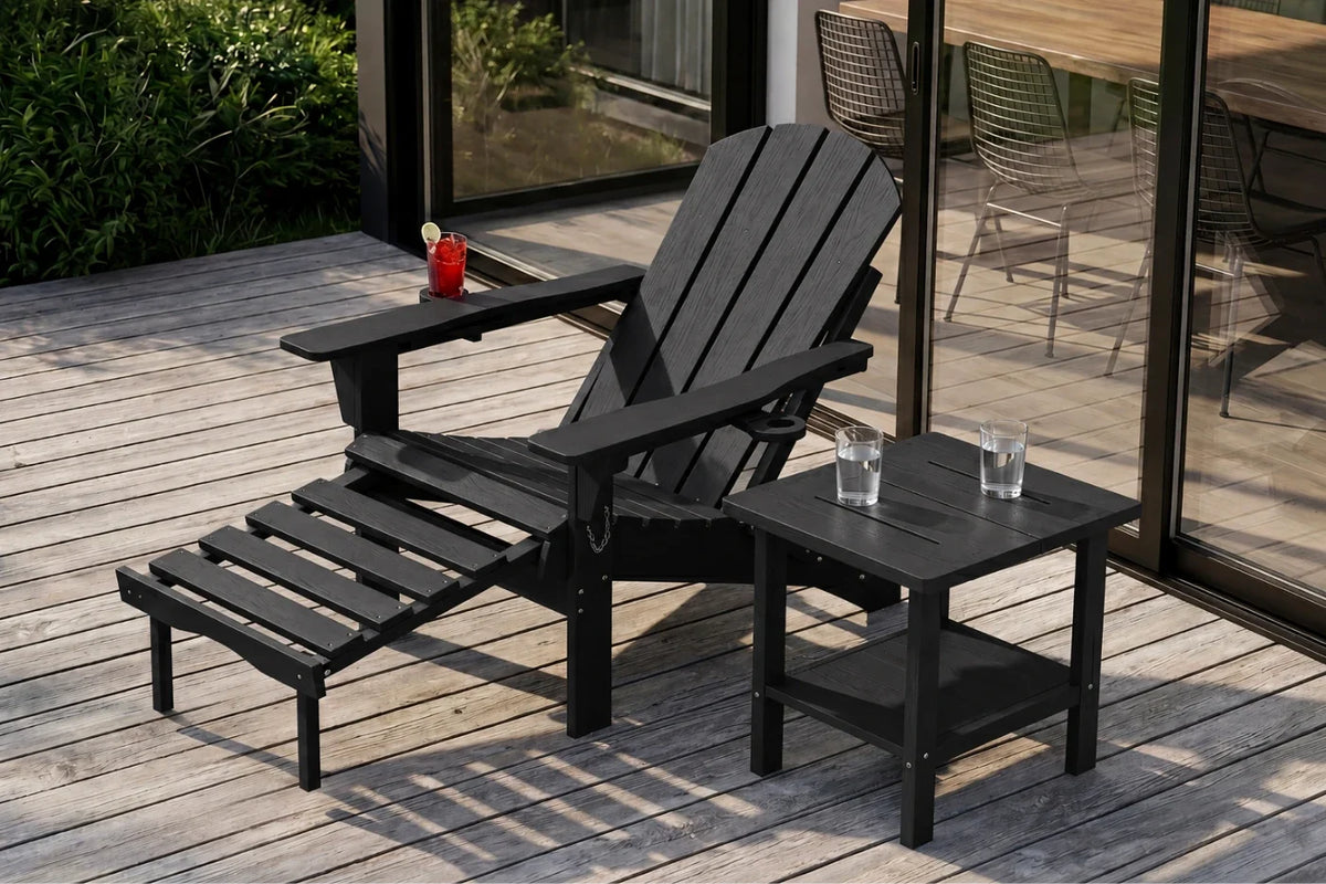 A black HDPE reclining adirondack chair with footrest, with a matching side table on a wooden deck. The scene is set in a patio environment with glass doors and greenery in the background.