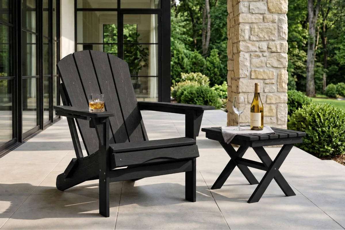 A black outdoor Adirondack chair sits on a stone patio beside a matching black side table with a bottle and glass, with a house exterior and greenery in the background.