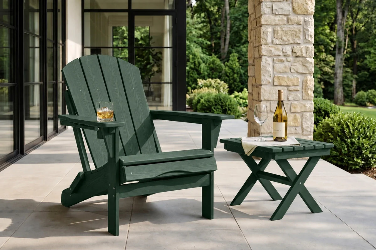 A dark green outdoor Adirondack chair sits on a patio beside a matching small side table, with a glass of whiskey and a wine bottle on the table. The patio is next to a stone pillar and large black-framed glass windows, with green trees and shrubs in the background.