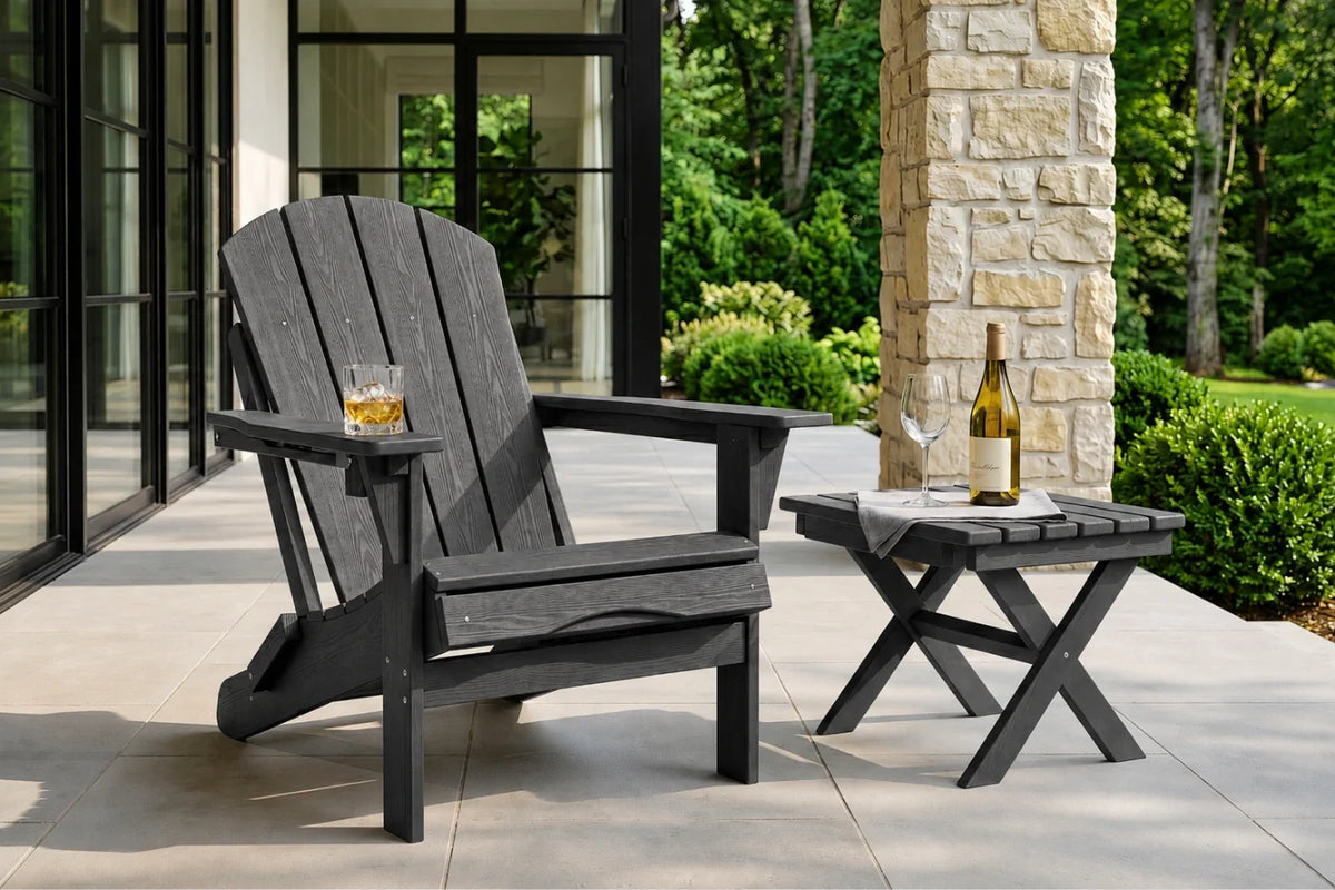 A dark gray outdoor Adirondack chair with a matching side table on a patio, with a glass of whiskey and a wine bottle on the chair armrest and table.