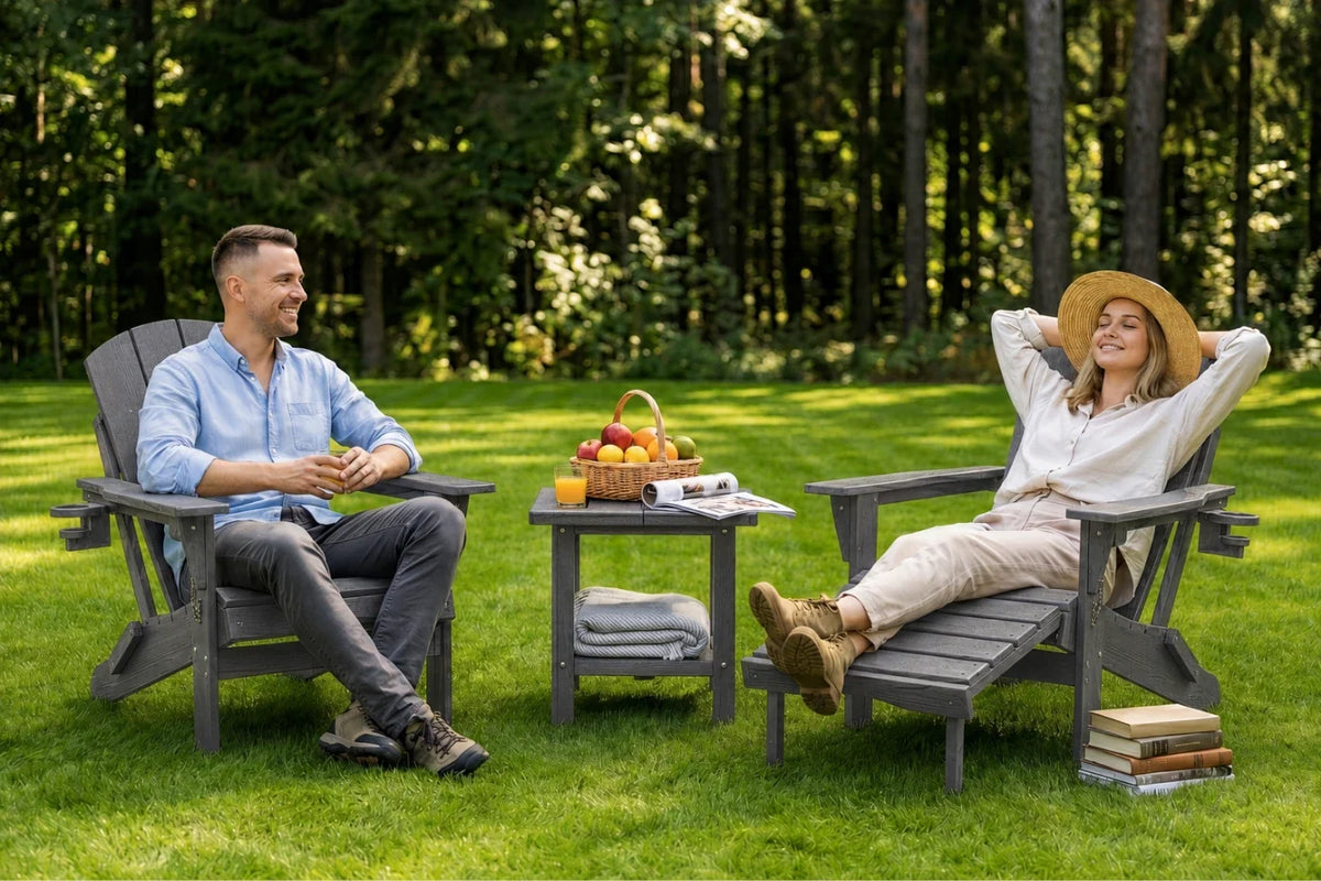A man and a woman relax in gray Adirondack chairs on a grassy lawn with a small table between them holding a fruit basket and a book. Trees fill the background.