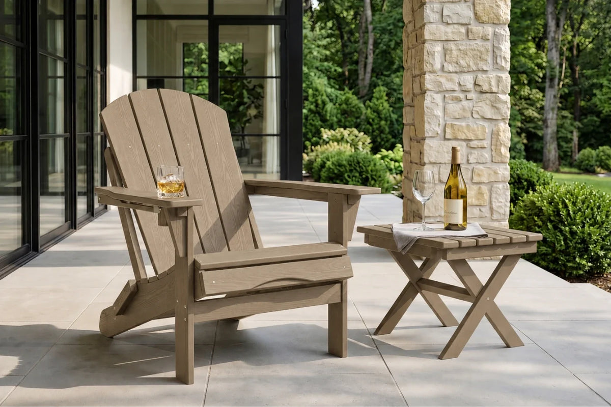 A light brown outdoor Adirondack chair sits on a patio beside a matching folding side table, with a glass of whiskey and a wine bottle on the table. The setting includes a stone pillar, green shrubs, and trees in the background.