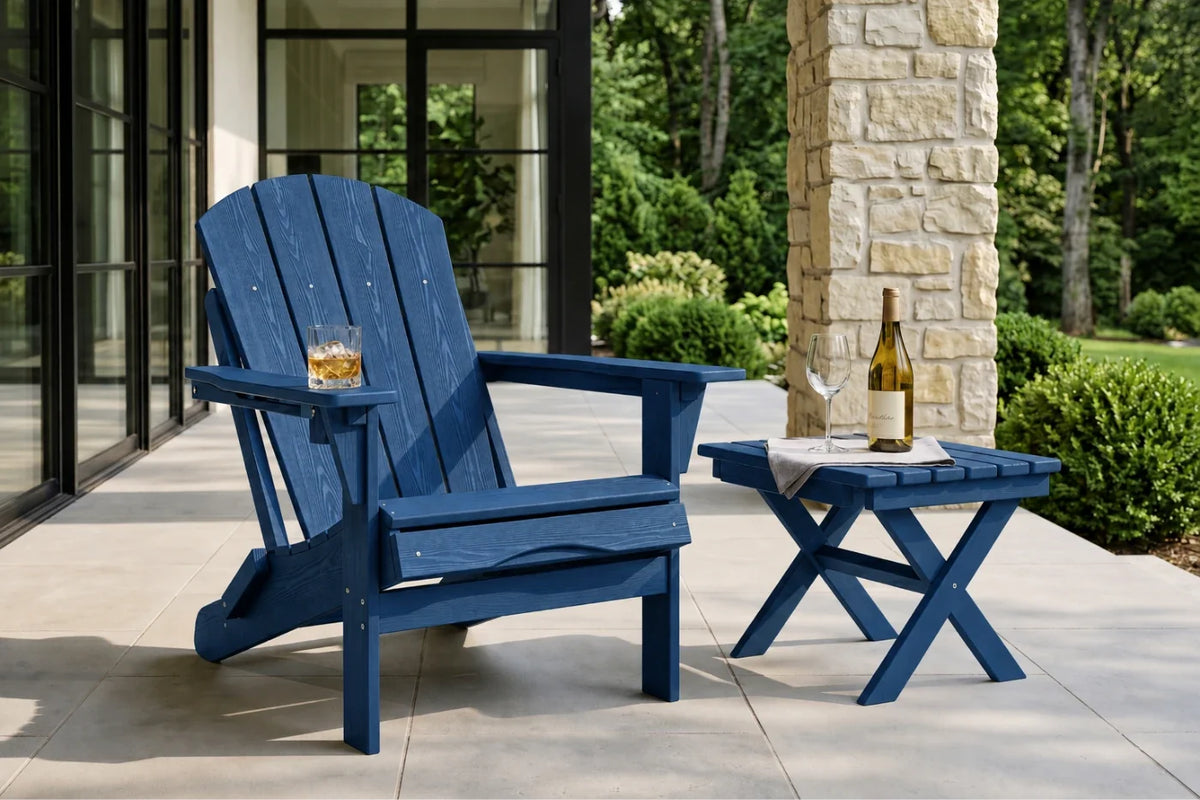 A navy blue outdoor Adirondack chair sits on a patio beside a matching blue side table with a wine bottle and glass, with a stone column and green garden in the background.