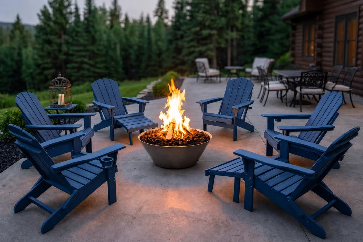 A set of navy blue Adirondack chairs arranged around a lit fire pit on a concrete patio, with a log cabin, trees, and outdoor furniture in the background at dusk.