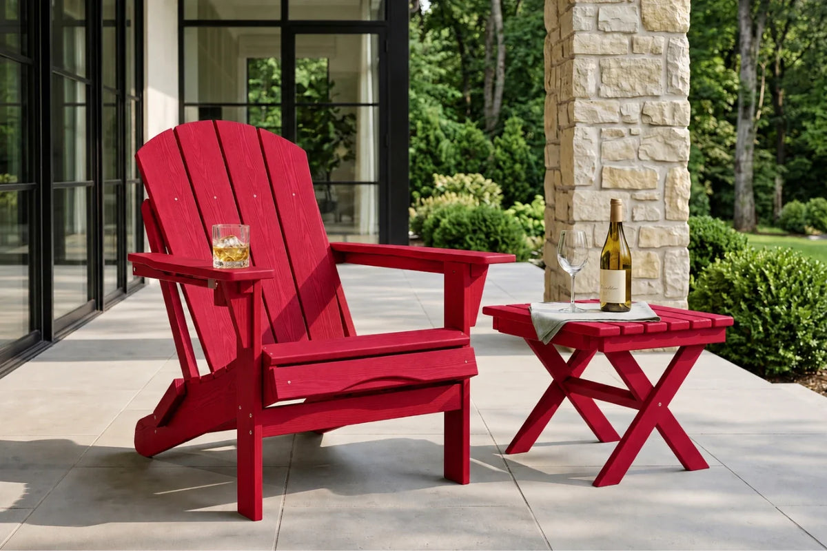 A red outdoor Adirondack chair sits on a patio beside a matching red side table with a wine bottle and glass, with a stone column and green garden in the background.