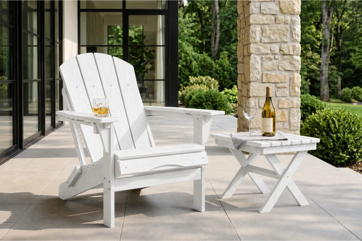 A white outdoor Adirondack chair with a matching side table on a patio, with a glass of whiskey in the chair's cup holder and a wine bottle on the table. The background shows a stone pillar, glass patio doors, and a lush green garden.