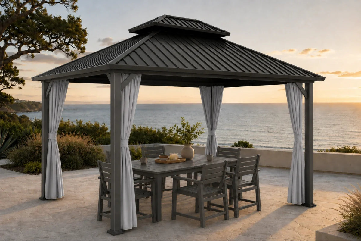 A dark gray hardtop gazebo with netting and curtains, set on a stone patio overlooking the ocean at sunset. A matching gray dining table and six chairs are placed underneath.