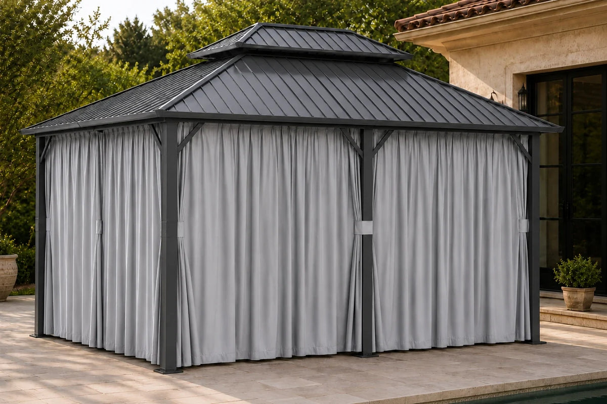 A dark gray hardtop gazebo with netting and curtains, situated on a tiled patio next to a house with green trees in the background.