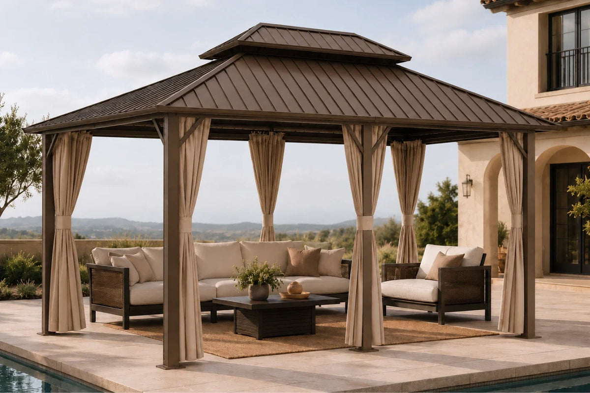 A brown hardtop gazebo with netting and curtains, furnished with a beige sectional sofa, two armchairs, and a coffee table, set on a stone patio next to a swimming pool with a house in the background.