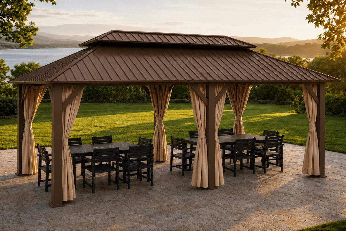 A brown hardtop gazebo with netting and curtains, furnished with a dining table and chairs, set on a stone patio with a green lawn and lake in the background.