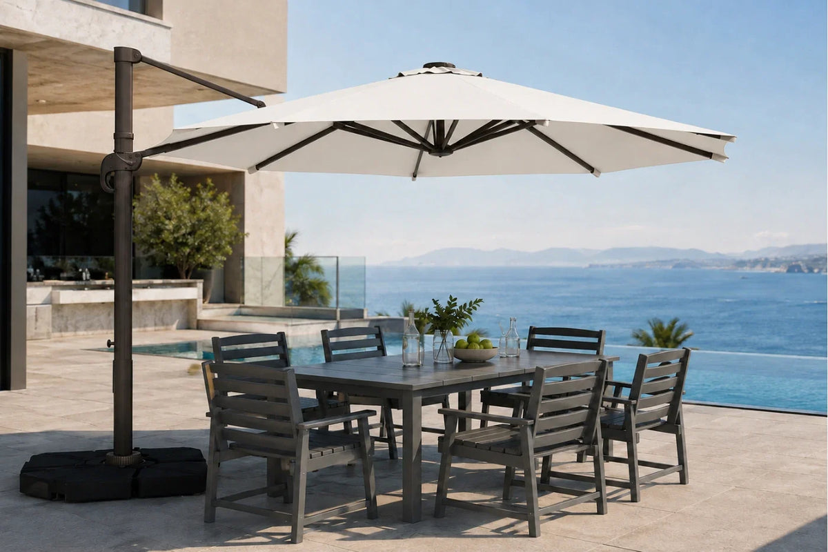 A large outdoor patio umbrella with a dark metal cantilever arm and black base, shading a rectangular gray dining table and six matching slatted chairs on a sunlit terrace overlooking a blue ocean and pool.