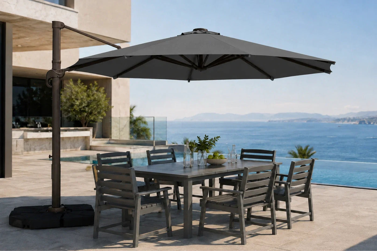 A large outdoor patio umbrella with a dark metal cantilever arm and black base, shading a rectangular gray dining table and six matching slatted chairs on a sunlit terrace overlooking a blue ocean and pool.