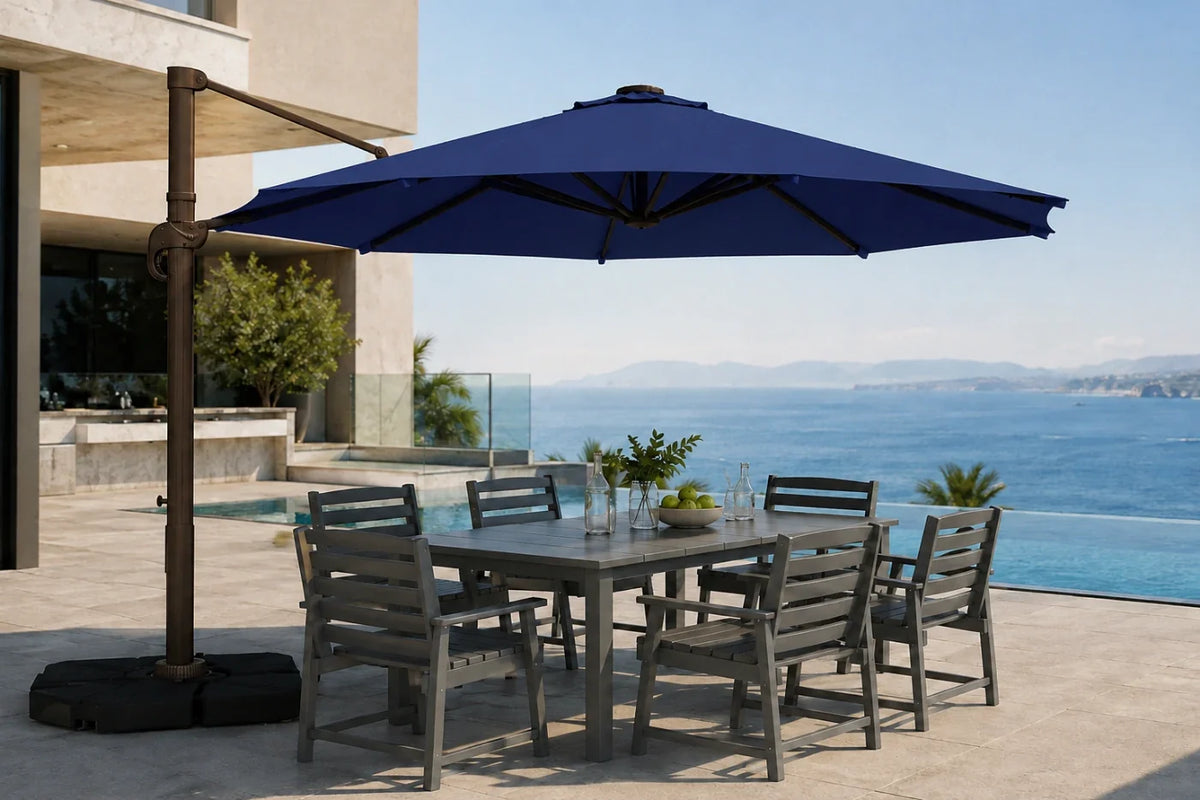 A large outdoor patio umbrella with a dark metal cantilever arm and black base, shading a rectangular gray dining table and six matching slatted chairs on a sunlit terrace overlooking a blue ocean and pool.