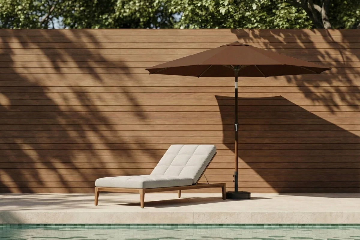 Chaise lounge with a brown umbrella by a poolside against a wooden wall.