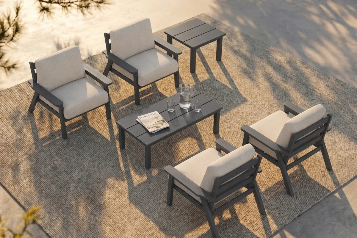 A 6-piece outdoor patio conversation set including four armchairs with creamy white cushions, one matching rectangular coffee table, and one matching side table, arranged on a textured beige rug with a concrete patio surface.