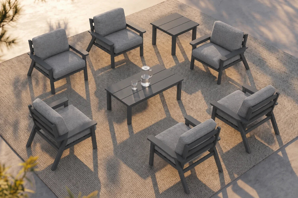 An 8-piece outdoor patio furniture set arranged on a woven rug, featuring six dark gray armchairs with gray cushions, one matching rectangular coffee table, and one matching side table.