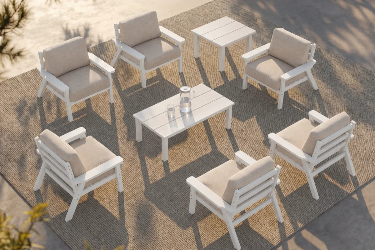 White outdoor patio furniture set with six armchairs, one matching rectangular coffee table, and one matching side table arranged on a woven rug. The chairs and tables have creamy white cushions, photographed from an elevated angle in a sunlit patio setting.