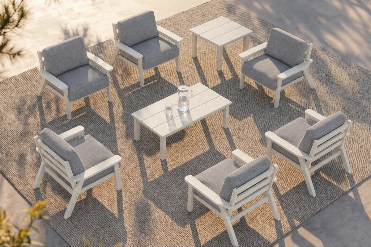 An outdoor patio conversation set with six white-framed armchairs with gray cushions arranged around one matching rectangular wooden coffee table and one matching side table on a woven rug.