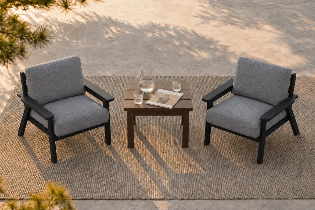 Two gray armchairs with black frames arranged around a small dark side table on a woven rug, photographed in an outdoor patio setting with dappled sunlight and shadows.