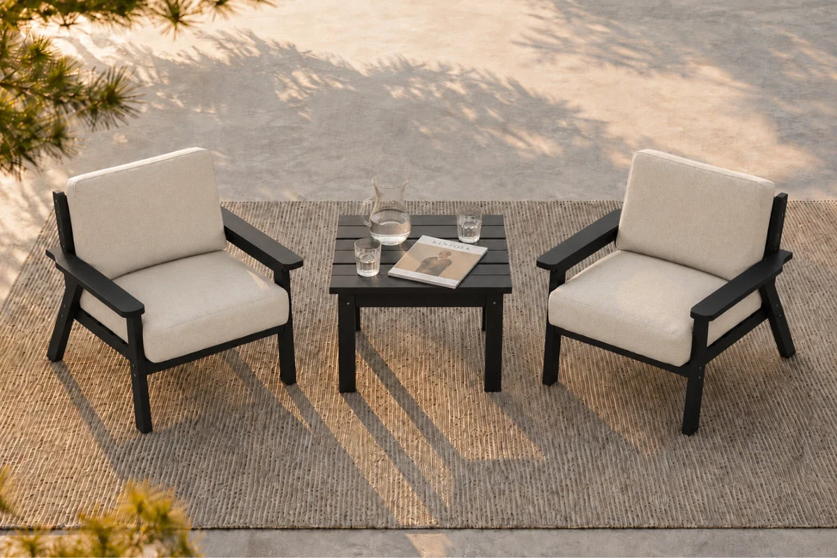 A 3-piece outdoor patio furniture set featuring two black-framed armchairs with creamy white cushions and a matching black slatted side table, arranged on a woven jute rug.