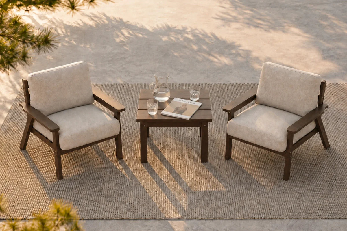 Two outdoor armchairs with creamy white cushions are arranged on a woven rug around a small square side table with a glass decanter and magazine, set on a concrete patio with dappled sunlight.