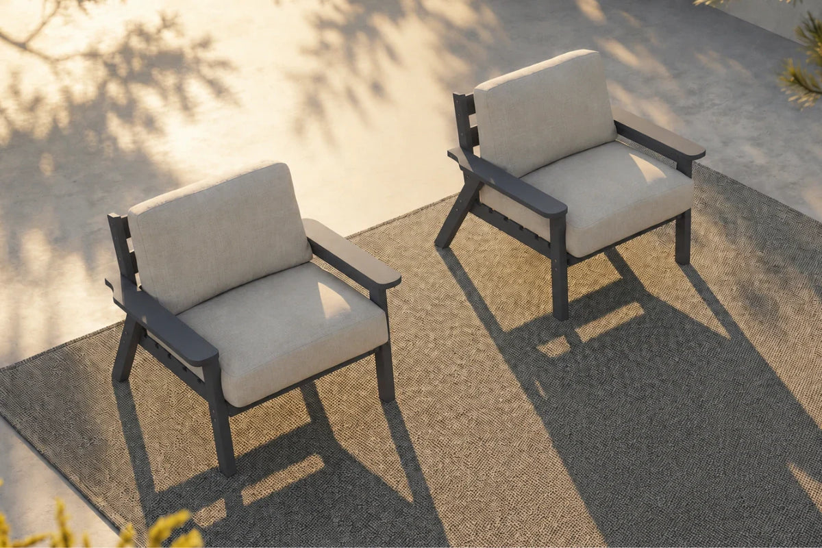 Two modern outdoor armchairs with creamy white cushions and dark gray frames, positioned on a textured gray patio rug with warm sunlight and leaf shadows.