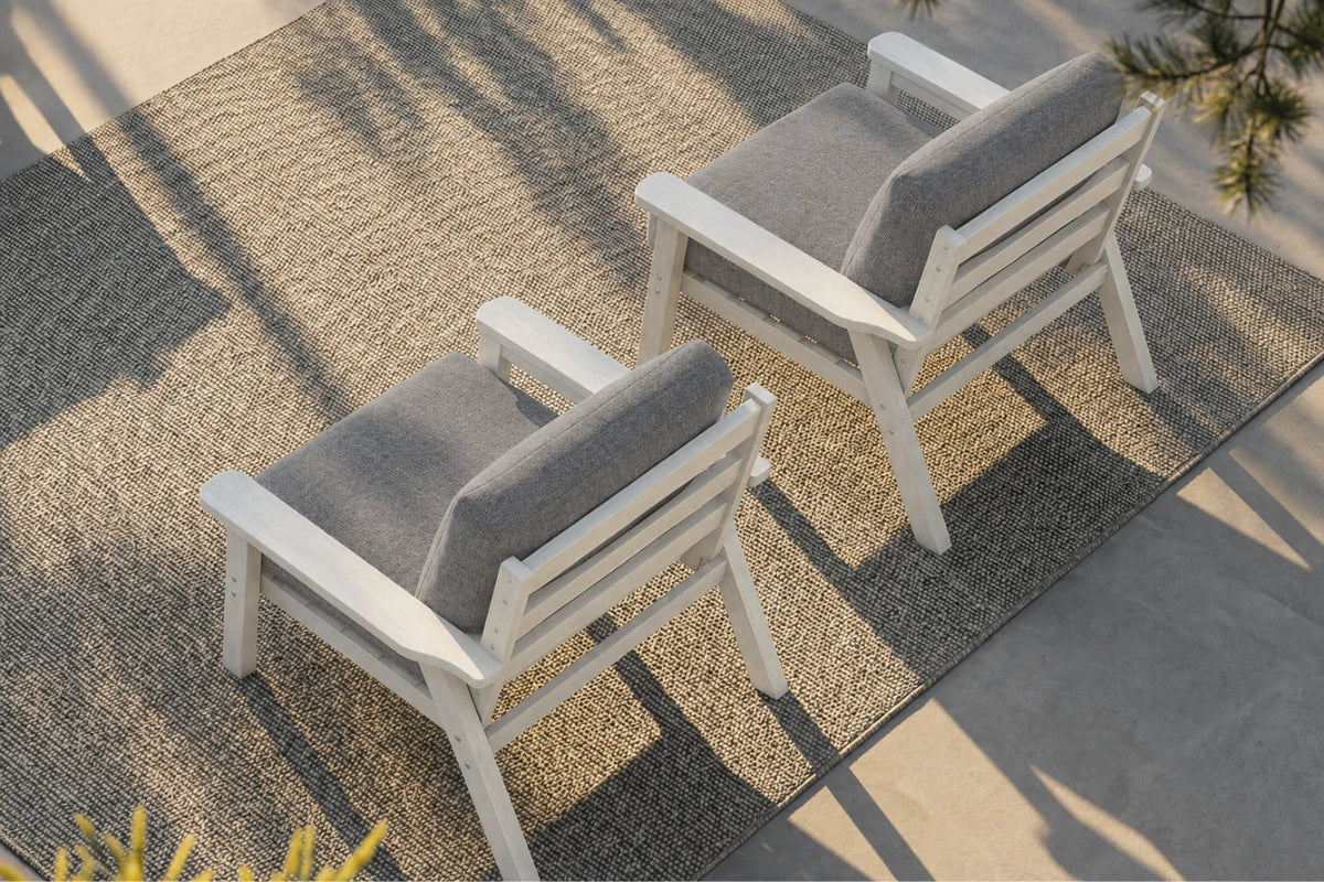 Two modern outdoor armchairs with white frames and gray cushions, arranged on a textured beige rug over a concrete patio.