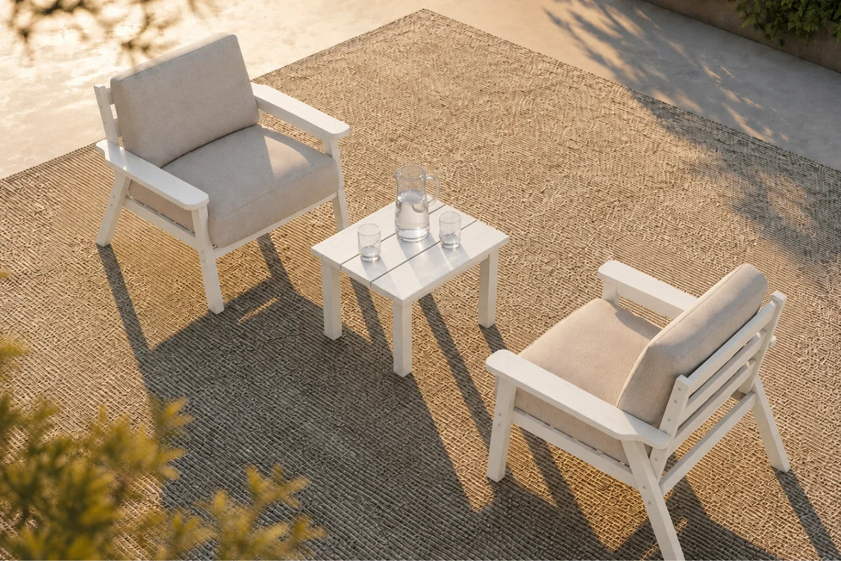 A high-angle view of a 3-piece outdoor patio furniture set consisting of two white armchairs with creamy white cushions and a small square white side table, arranged on a woven brown rug.