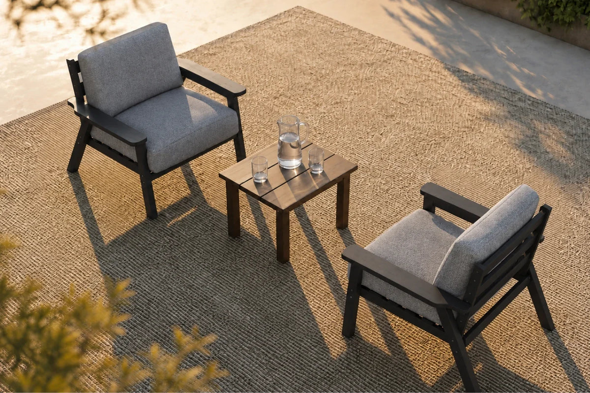 Two outdoor armchairs with gray cushions and black frames, arranged around a small square black side table on a woven rug in a patio setting.