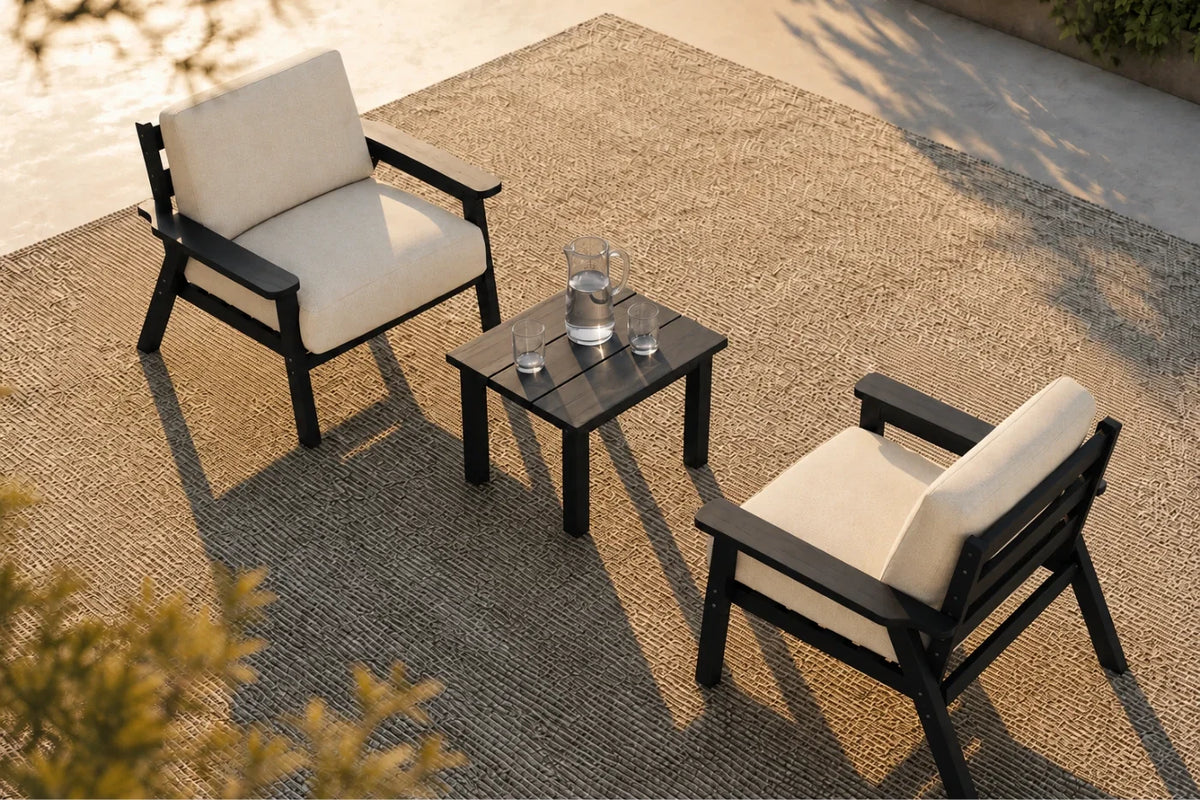 Two outdoor armchairs with cream cushions and black frames arranged around a small black side table on a woven patio rug, photographed from above in warm sunlight.