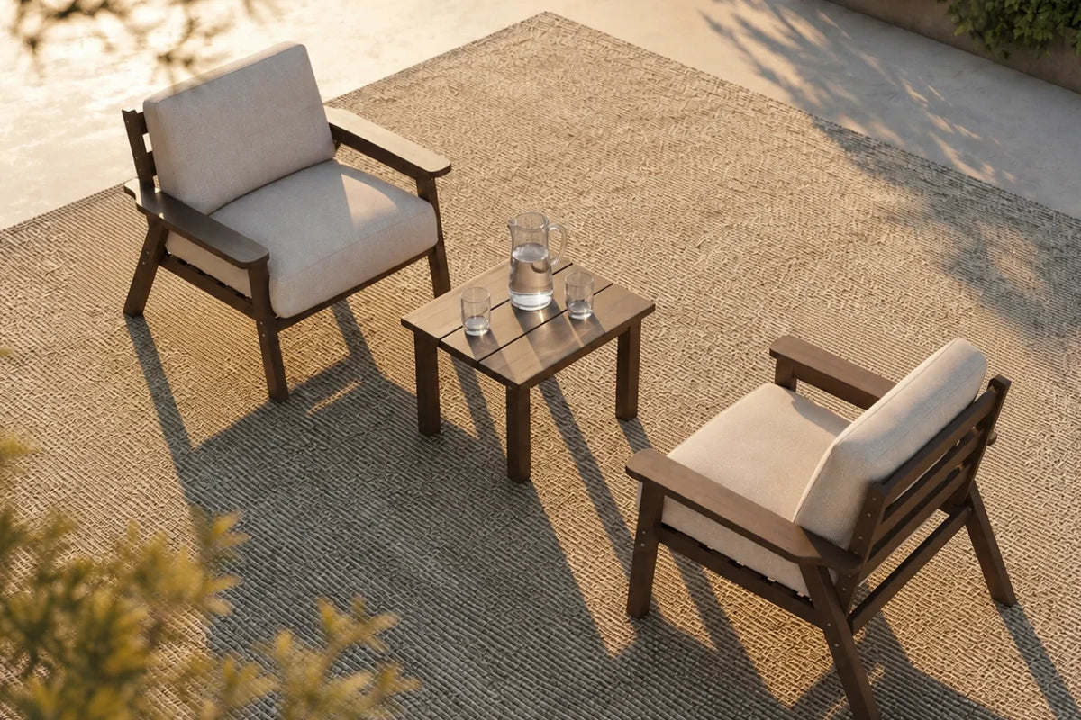 Two outdoor armchairs with creamy white cushions and brown frames are arranged around a small square brown side table on a woven patio rug, with warm sunlight casting long shadows.