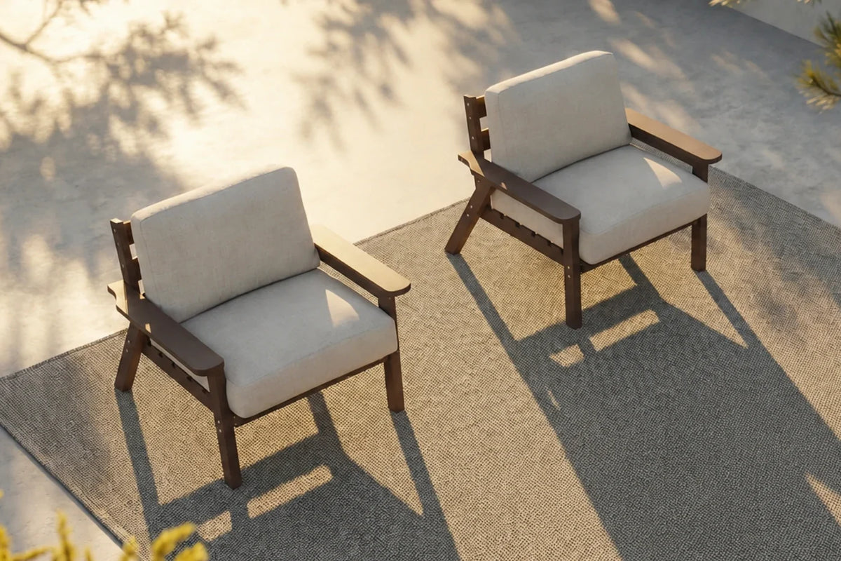 Two modern outdoor armchairs with creamy white cushions and brown frames, arranged on a textured gray patio rug under warm sunlight with leaf shadows.