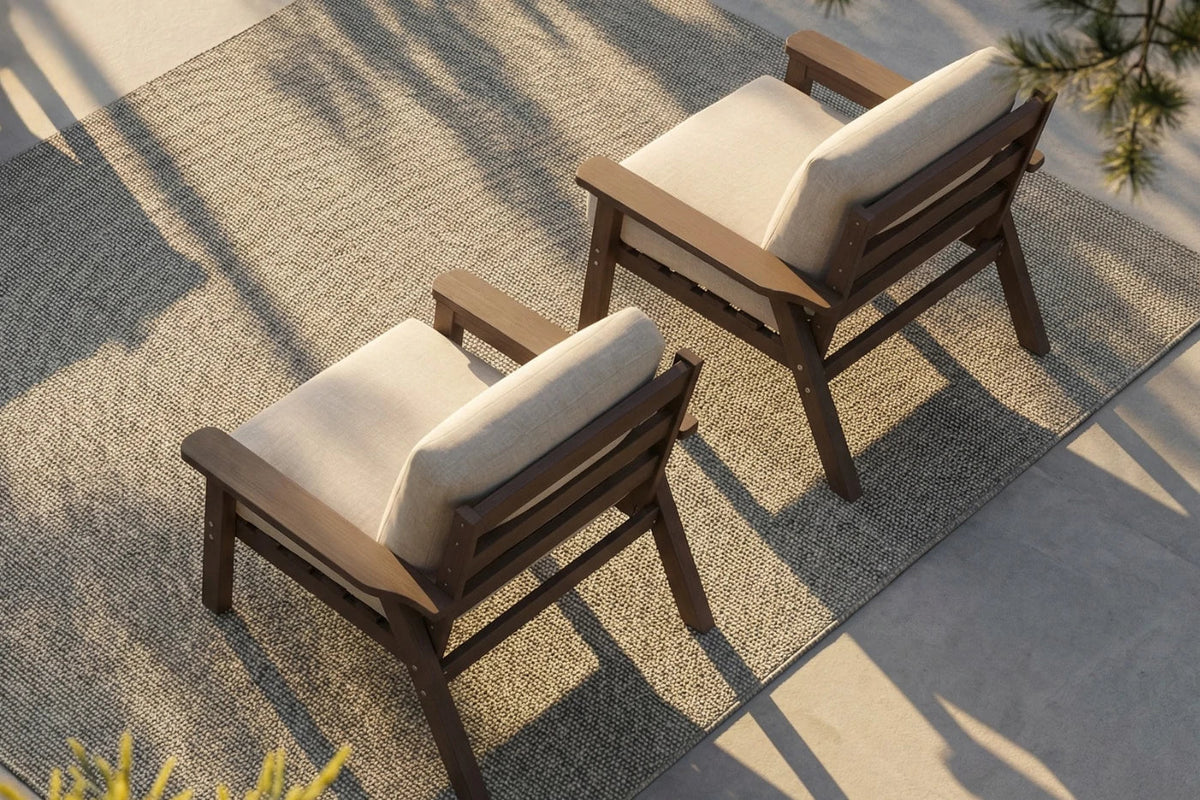 Two outdoor armchairs with brown wooden frames and creamy white cushions, arranged on a textured beige rug over a concrete patio with dappled sunlight.