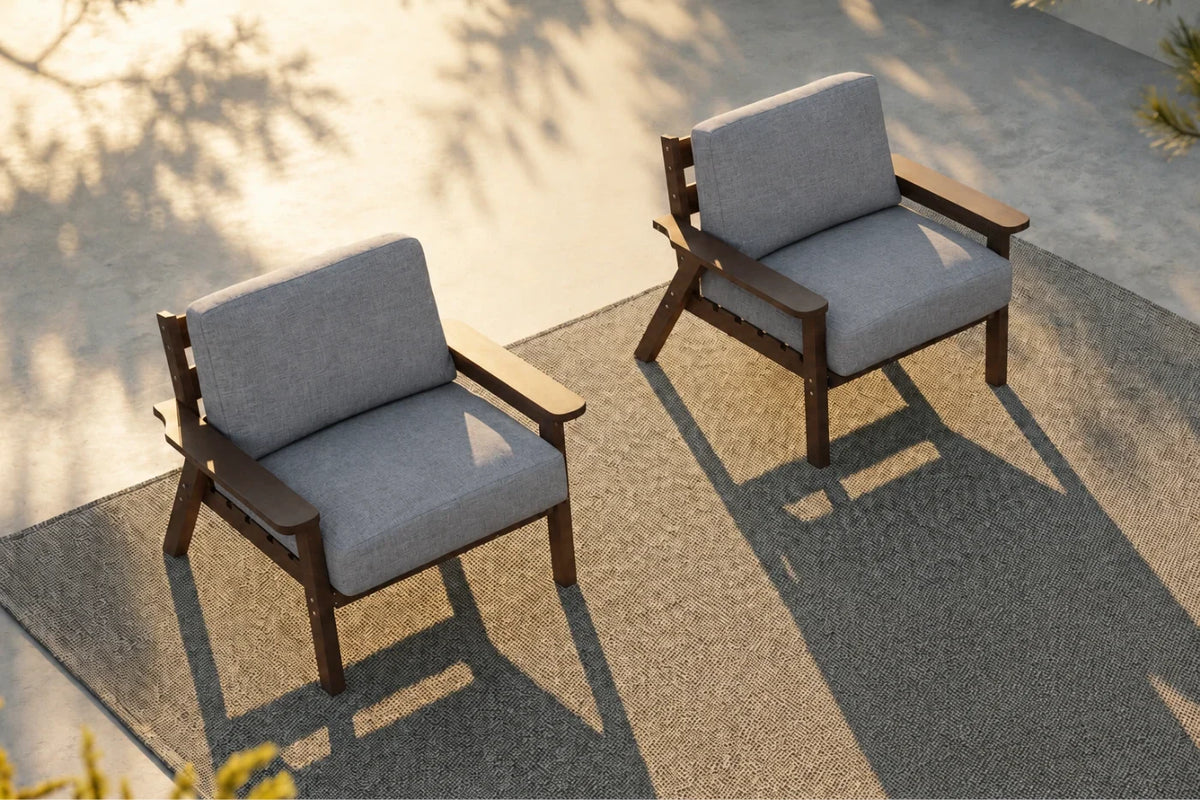 Two modern outdoor armchairs with gray textured cushions and brown wooden frames, positioned on a textured concrete patio with warm sunlight and leaf shadows.