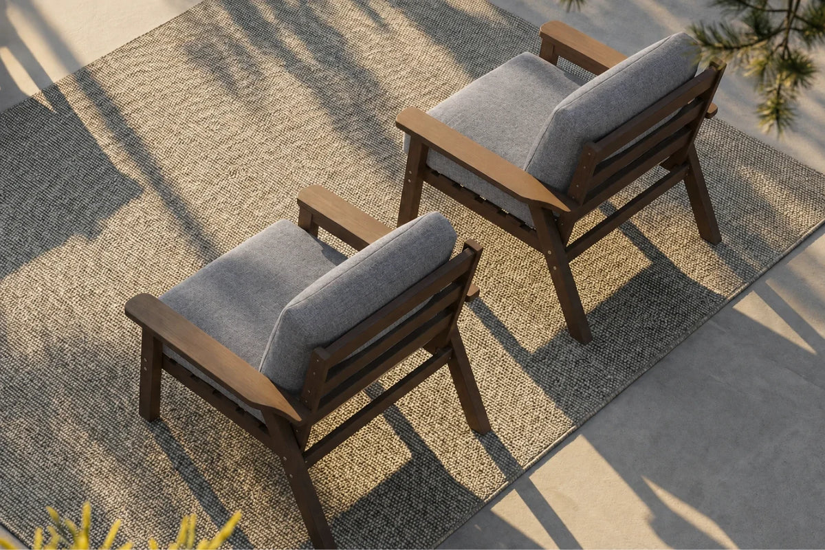 Two outdoor armchairs with brown wooden frames and gray cushions, arranged on a textured beige rug in a patio setting.